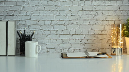Stylish workplace with opened notebook, pencil holder, coffee cup and books on white table against brick wall