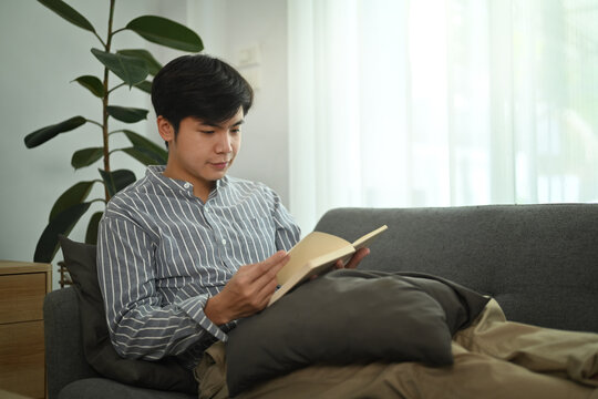 Calm Millennial Asian Man Relaxing Co Couch And Reading Literature Story Book In Bright Living Room. Domestic Lifestyle Concept