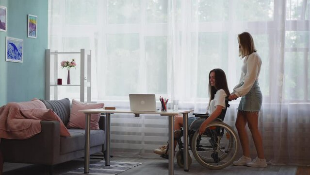 Woman pushes girl friend with disability in wheelchair with muscular dystrophy. Caring person helps freelancer get to workplace to use laptop in apartment