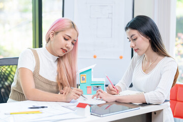 Fototapeta premium Concept of engineering consulting, Two female engineers discussing about model of building together; Two female architects are studying blueprint of building house.