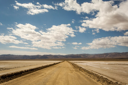 PANAMINT VALLEY, CA, USA. A long, desolate dirt road extends into a distant horizon under blue, cloudy skies.