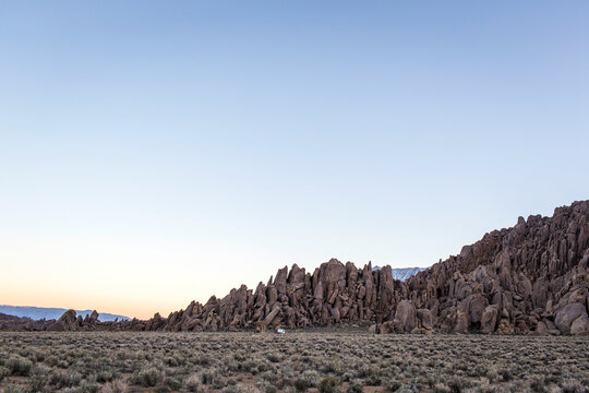 Lone Pine, CA, USA. A lone white camper van looks tiny against a jumble of boulders, a flat sage valley floor and an empty sky at dusk.