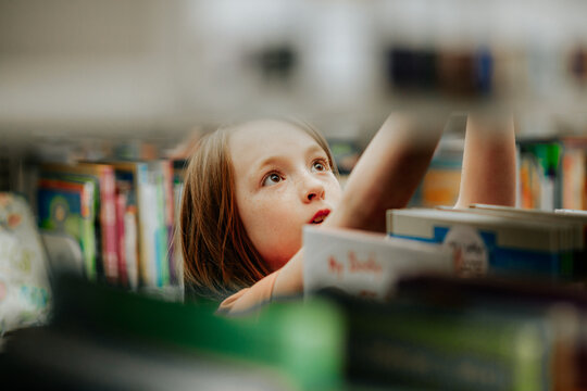 Young Girl Reaching For Library Book On Shelf