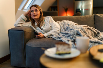 A woman in her pajamas is laying on the couch in the living room, she is covered with a blanket and is enjoying her day off and watching movies holding a TV remote in her hand - obrazy, fototapety, plakaty