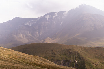 Obraz premium Majestic mountain landscape - mountain peak with glacier in soft light white pastel mist, golden sunbeams in sunny and cloudy day, yellow slopes with dry alpine meadow in autumn. Joyful landscape.