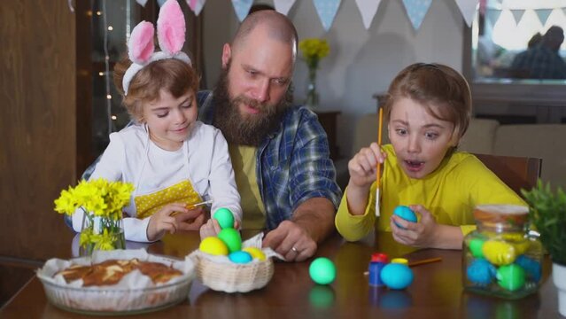 Easter Family traditions. Father and two caucasian happy children with bunny ears dye and decorate eggs with paints for holidays while sitting together at home table. 