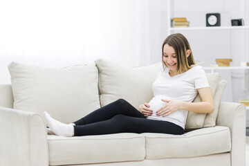 Obraz premium Smiling regnant woman put her hands on her stomach on white sofa.