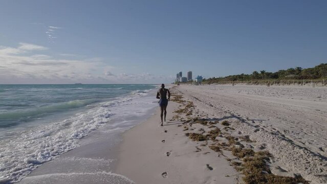 Tracking aerial footage of muscular male athlete running down a shoreline with cityscape in distance