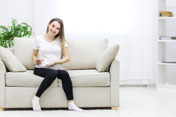 Smiling pregnant woman sitting on white sofa with two small socks.