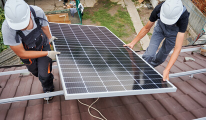 Men mounters lifting up photovoltaic solar moduls on roof of house. Electricians in helmets...