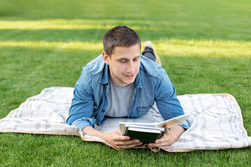 Young man relaxing and reading book while lying on lawn. Happy Man Lying On Green Grass and study remotely. Adult student reading in the park. Enjoying student life. smiling man working on outdoor	