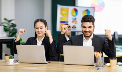 Millennial Asian Indian professional successful bearded male businessman and female businesswoman employee colleague in formal business suit sitting holding fists up celebrating job deal achievement