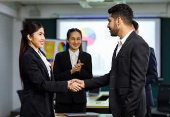 Millennial Asian Indian professional bearded male businessman shaking hands done deal female businesswoman in formal business suit while staff employee colleagues standing clapping hands celebrating