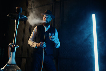 Front view of resting man smoking hookah at home workplace with neon lighting