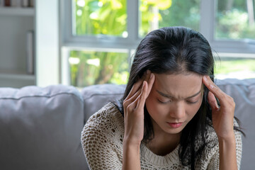 Headache. Young Asian woman is sitting on a sofa with her eyes closed, touching her head while suffering from a migraine.