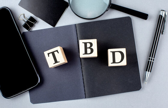 Word TBD on wooden block on black notebook with smartpone, credit card and magnifier