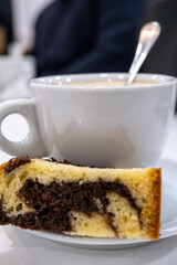 Café con leche en una taza blanca de porcelana con la cucharilla de metal dentro y un trozo de bizcocho con chocolate apoyado en el plato.
