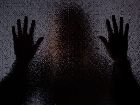 Blurred silhouette of a mysterious woman standing behind frosted glass with both hands against the glass. Concept of asking for help, loneliness or depression