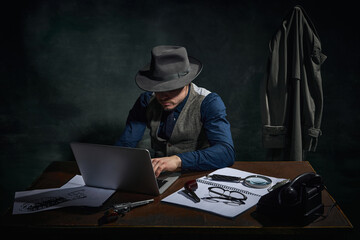 Professional detective in fedora hat sitting at table and working on laptop over dark green vintage...