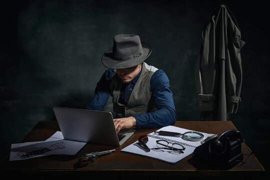 Professional detective in fedora hat sitting at table and working on laptop over dark green vintage background. Professional investigation. Concept of occupation, character, history. Retro style
