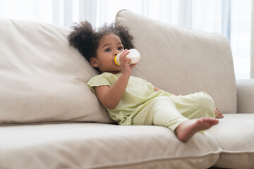 Cute African American little child girl with milk bottle. African American little girl eating milk from nipples bottle while playing on sofa at home