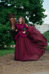 Obraz na płótnie Canvas Beautiful elegant woman in long flowing dress and thoroughbred brown horse. Walk with horse in woods. Aristocratic young lady holding long vintage dress and well-bred stallion. Traditional horse farm