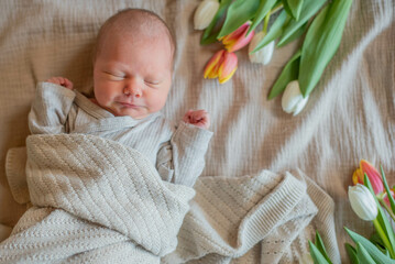 smiling newborn baby girl lying on a beige blanket in a beige bodysuit surrounded by white and pink...