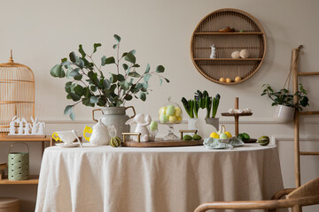 Warm and spring dining room interior with easter accessories, shelf on wall, round table, vase with green leaves, colorful eggs, rabbit sculpture and personal accessories. Home decor. Template.