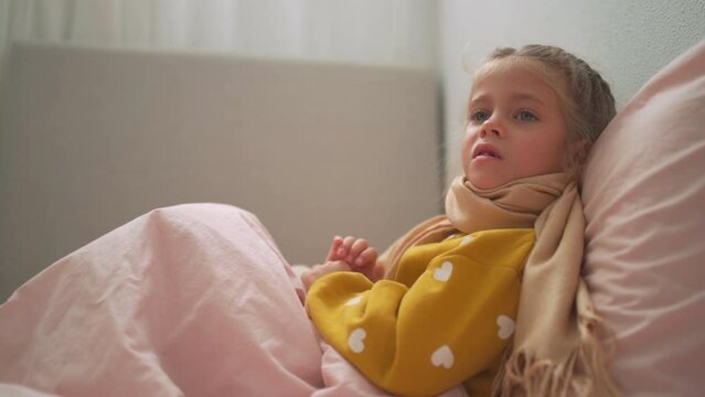 Child girl in plaid and scarf suffering from Coronavirus or allergy, sneezes snot into napkin on sofa at home 