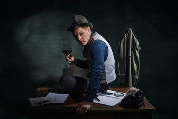 Professional detective in fedora hat and classical clothes sitting on table with glass of red wine over dark green vintage background. Concept of occupation, character, history. Retro style