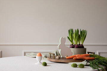 Creative composition of dinning room interior with copy space, round table, wooden trace, orange carrot, hyacinth in pot, hare sculpture, eggs and personal accessories. Home decor. Template.