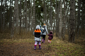 Back of mom and children with backpacks walking along the forest road after rain together.