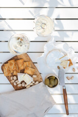 Two glasses of white wine and a wooden plate with cheese and nuts on a white table outdoors.