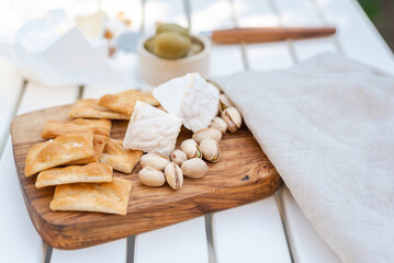 Wine appetizers namely pistachios, camembert cheese, saltine crackers and olives on a white table outdoors.