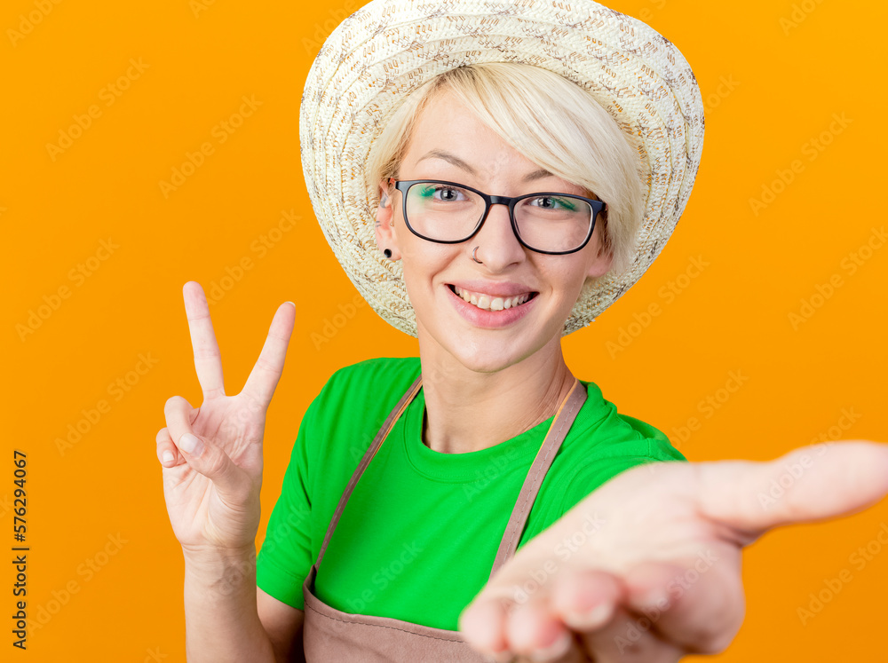 Canvas Prints young gardener woman with short hair in apron and hat looking at camera smiling cheerfully showing v-sign standing over orange background