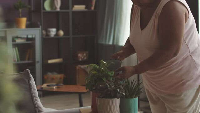 Medium shot of mature African American woman listening to music in headphones while watering potted house plants in cozy living room