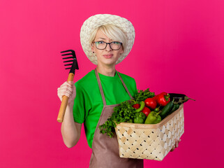 young gardener woman with short hair in apron and hat holding crate full of vegetablest and mini...