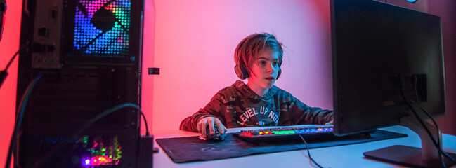 Image of immersed teenage gamer boy playing video games on computer in dark room wearing headphones and using backlit colorful keyboard © stenkovlad