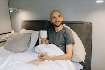 A man in bed reads a book and drinks hot tea or coffee and thinks about something