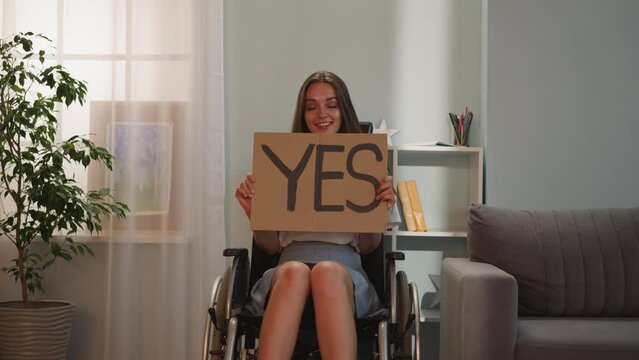 Cheerful woman with disability turns poster with word Yes to camera with smile on face. Young person with muscular dystrophy sits in wheelchair demanding tolerance