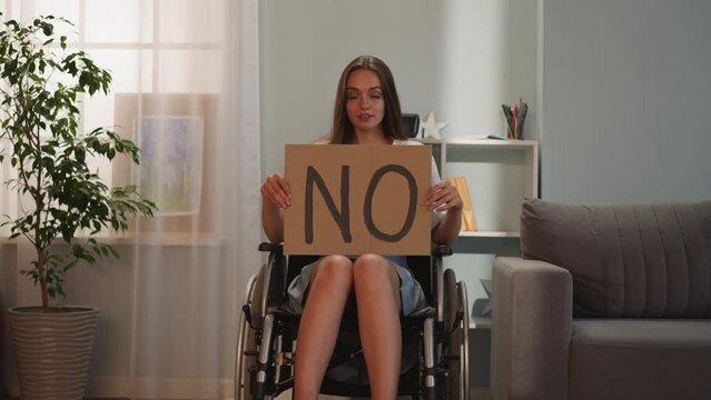 Woman with disability shows poster with word No looking in camera. Young female with paralysis protests against social inequality sitting in wheelchair at home