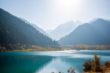 Autumn landscape. Tall old firs grow on the slopes of the mountains surrounding the mountain lake. A beautiful view of high mountains in a blue haze and a lake with azure water.