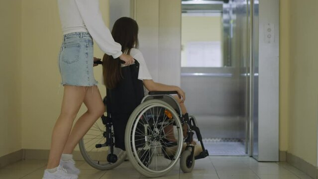 Woman walks in elevator cabin pushing wheelchair with sister with paralysis after door opened. Female person helps family member with disability get to apartment