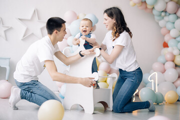 Happy parents have fun and play with their son celebrating first birthday among colorful balloons.