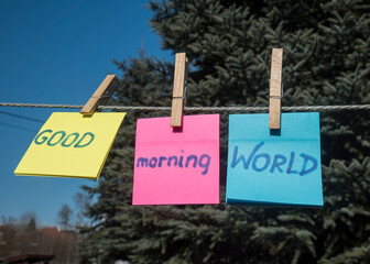 Napis GOOD MORNING WORLD na kolorowych kartkach powieszonych na klipsach na sznurku, na tle iglastych drzew w słoneczny dzień.
