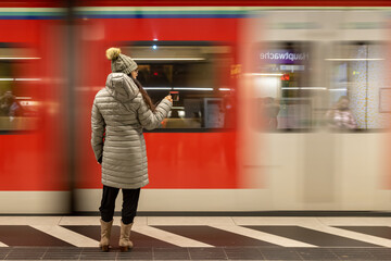 mulher com roupa de inverno na plataforma do metro segurando uma xicara de chá em quanto o trem passa borrado ao fundo  - obrazy, fototapety, plakaty