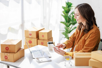 Concept of parcel delivery and selling online,Female seller or retailer using smartphone to check parcel box before delivery and using laptop checking day's sales on the online store