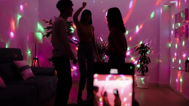 Three young friends dancing and singing at a private party in the living room at home and broadcasting live for their followers on their social media channels with a mobile phone