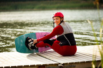 Woman in wetsuit, helmet and life vest sitting with wakeboard on a wooden pier. Sunny summer day. Safety in sport. Water sports in Finland. Insurance concept