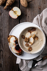 Bowl of homemade Jerusalem artichoke and apple cream soup and toast with homemade pate with fresh apple and cherry jam and fresh sun choke tubers and apple on wooden table.